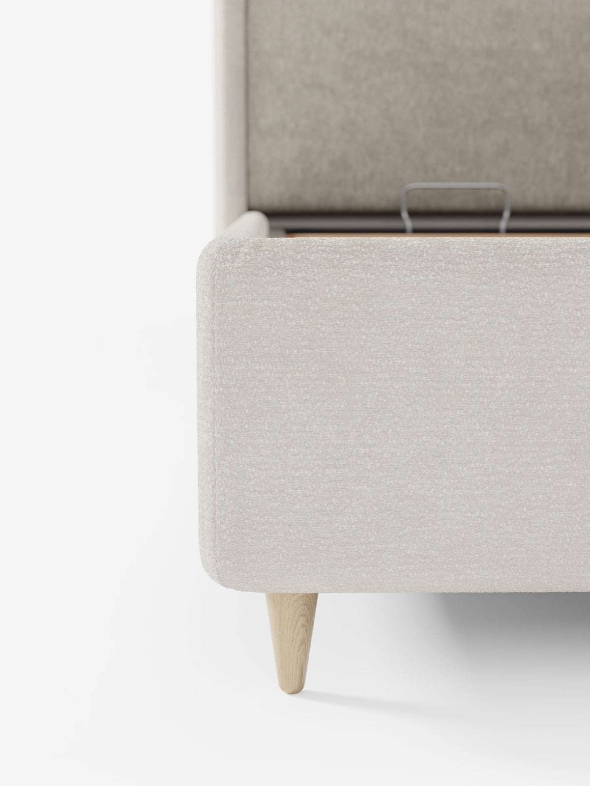 Un primo piano di un letto imbottito VALKIRIA bicolore color beige con contenitore - cuscini dello schienale e testiera curva avvolgente con gambe affusolate in legno chiaro, che mostra lo spazio di contenimento aperto, la maniglia in metallo e la testiera curva grigio chiaro.