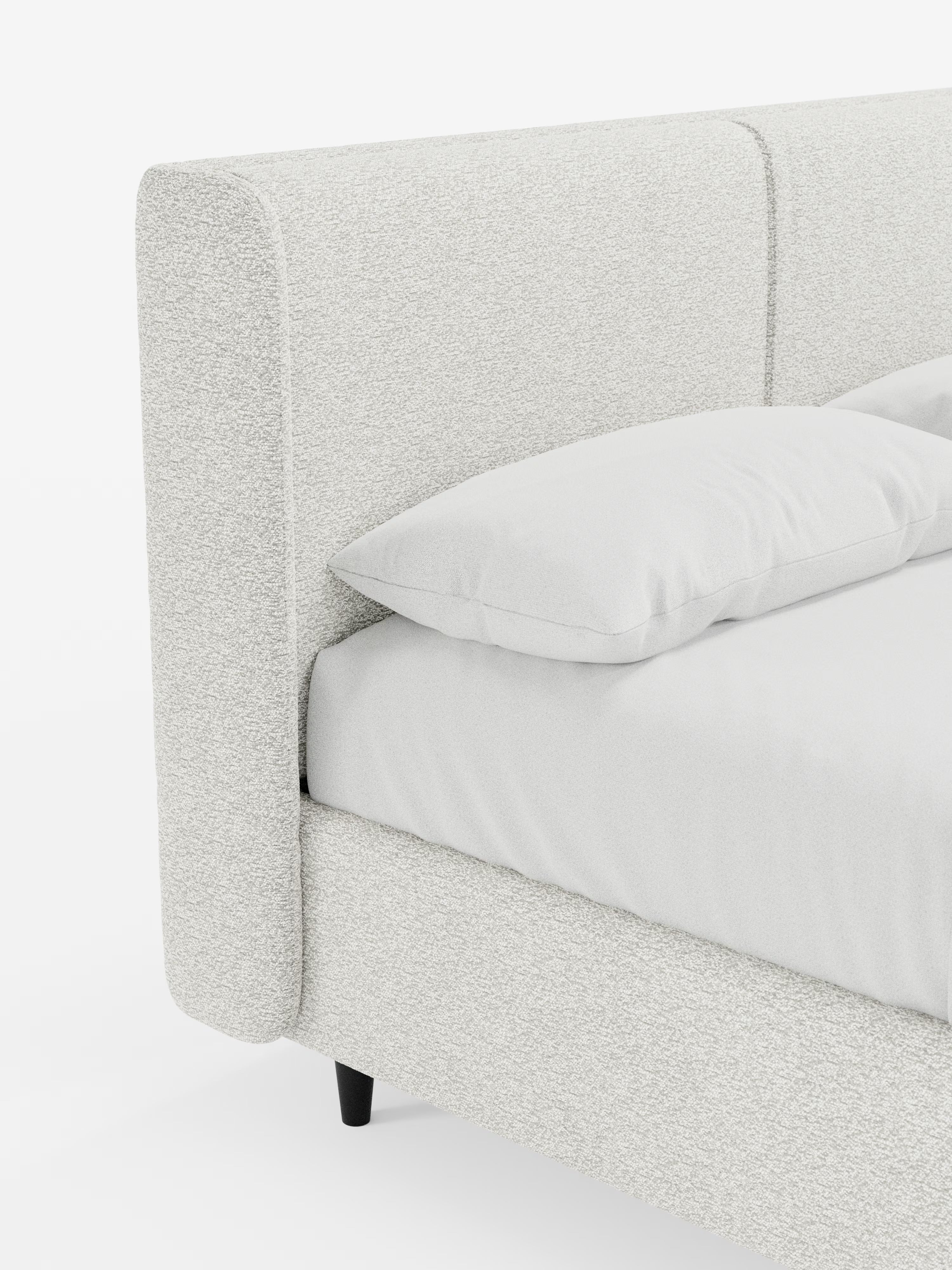 Primo piano del letto di design VALKIRIA di Letti.store, un letto imbottito rivestito in tessuto bianco sporco testurizzato certificato Oeko-Tex®. Questo letto moderno si distingue per la testiera alta e avvolgente, con due cuscini imbottiti abbinati, e per il profilo ribassato che ne esalta il design contemporaneo. Sono visibili un cuscino e un materasso coordinati con un lenzuolo con angoli. Il letto poggia su gambe corte e nere su uno sfondo bianco a tinta unita ed è dotato di un pratico vano