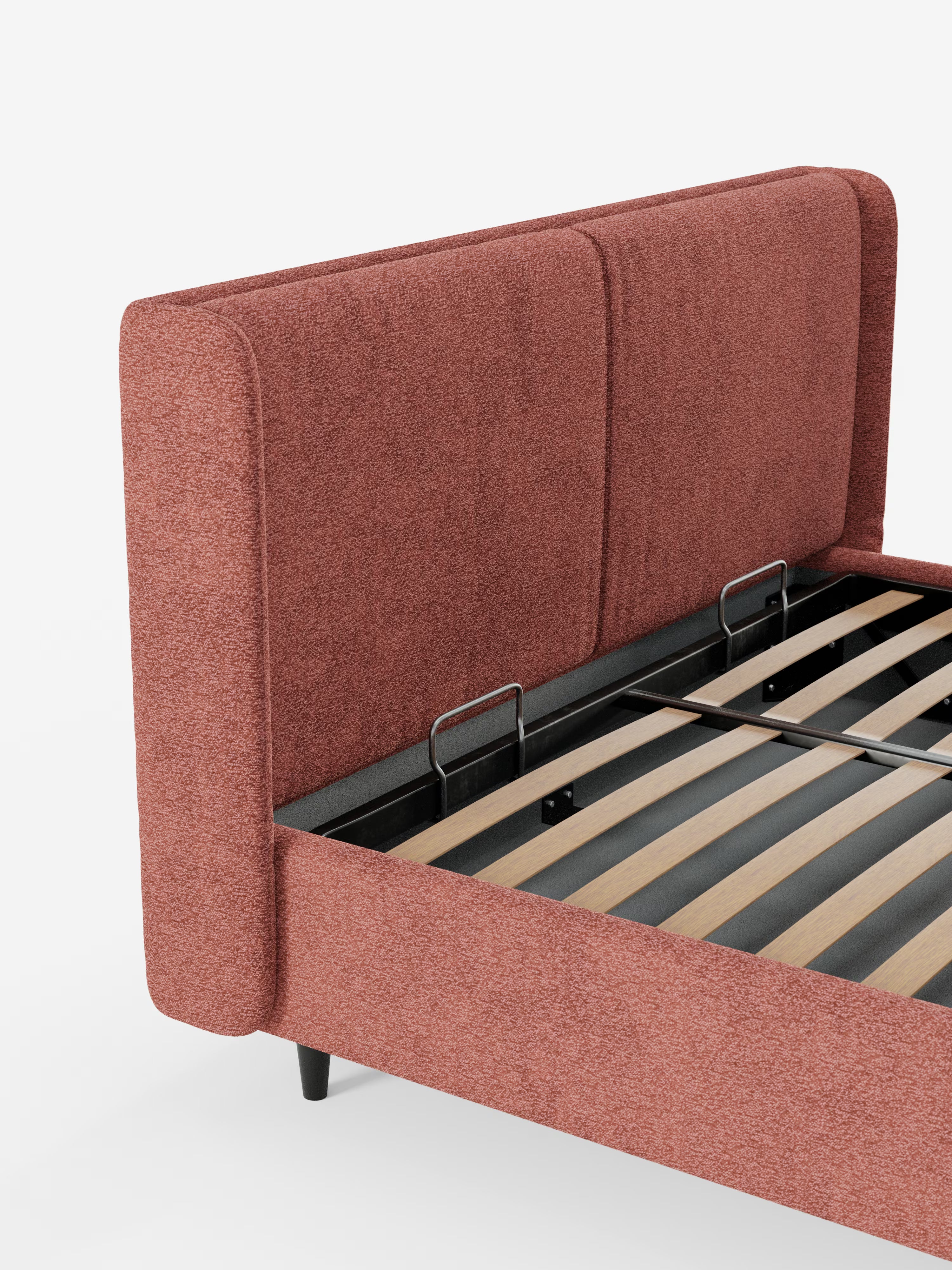 Qui viene presentato il letto di design VALKIRIA con vano contenitore e testiera avvolgente di Letti.store, caratterizzato da una testiera e da un lato imbottiti color ruggine con tessuto Oeko-Tex®. Questo design contemporaneo mette in risalto le doghe in legno a vista e i dettagli della struttura in metallo nero, che risaltano sullo sfondo bianco. È visibile solo una parte del letto imbottito, senza materasso; due cuscini imbottiti adornano la caratteristica testiera avvolgente.