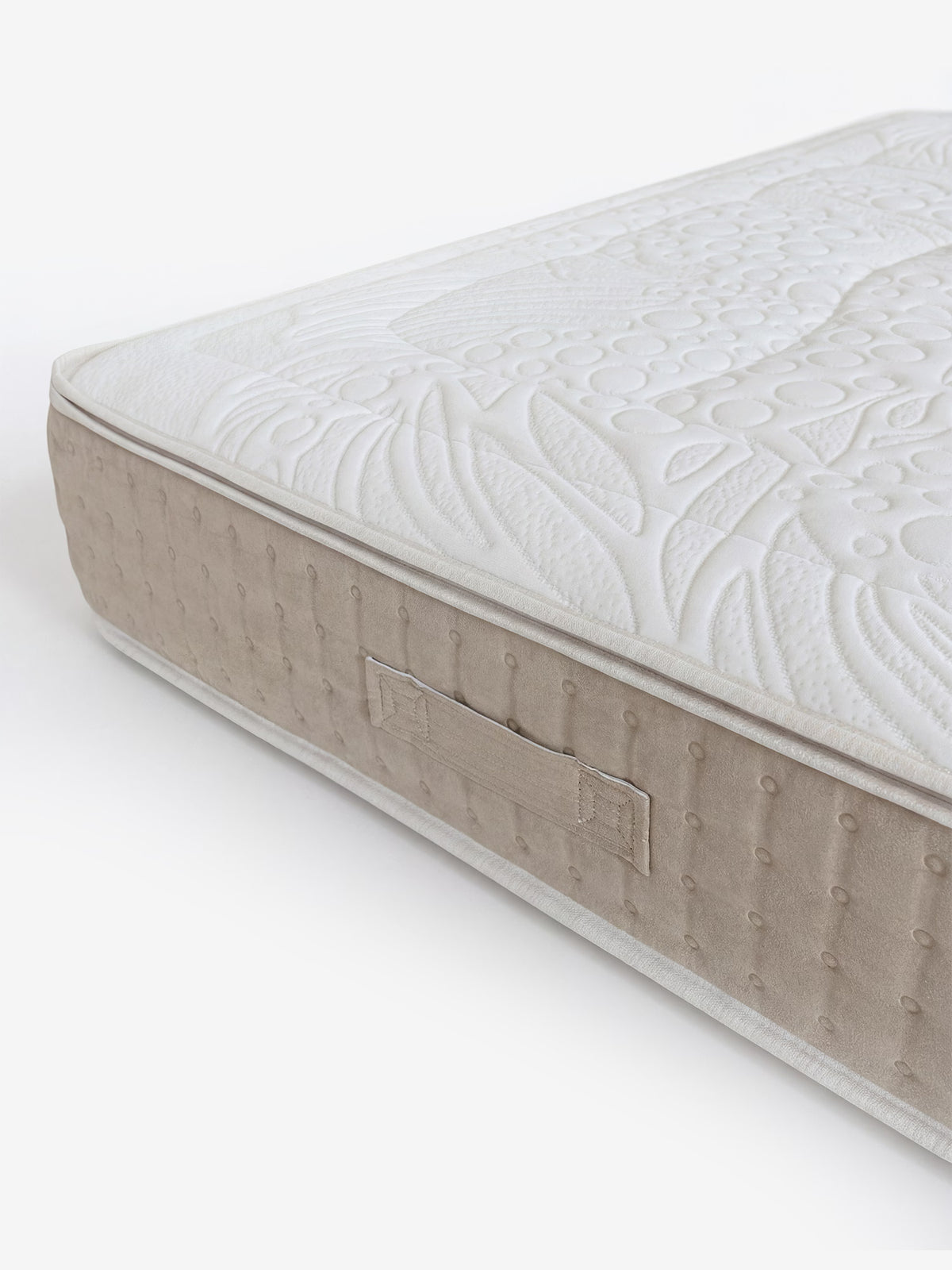 Un primo piano di un angolo del materasso beige e bianco mette in risalto un pannello laterale testurizzato con piccoli puntini, una maniglia in tessuto e una superficie superiore trapuntata caratterizzata da un intricato motivo floreale e geometrico. Il VELTHA – Materasso Bio-Adattivo con molle insacchettate e Lattice Naturale a 7 zone di Divani.Store è dotato di 800 micro molle insacchettate per garantire un comfort ottimale, su uno sfondo bianco a tinta unita.