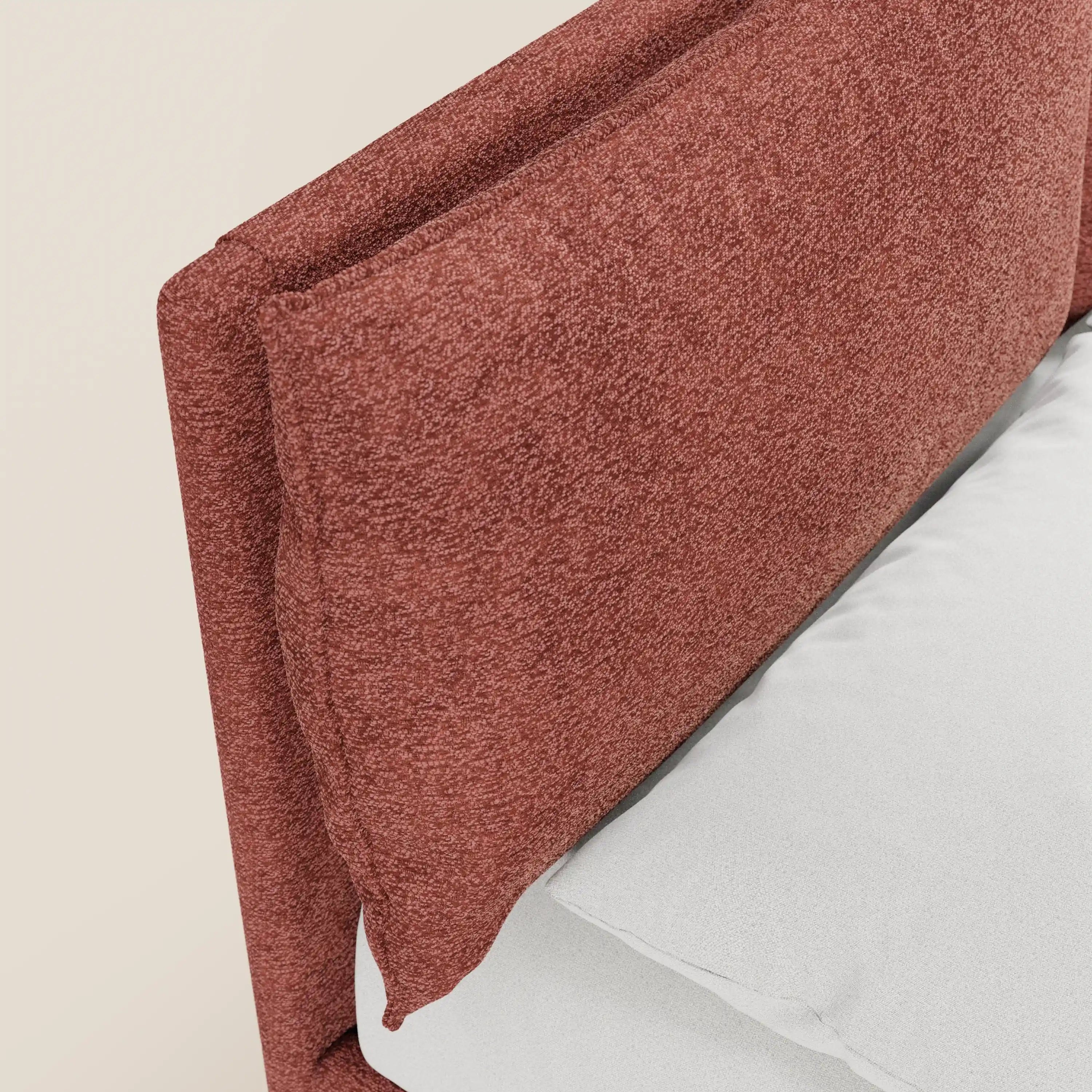 Primo piano del letto imbottito ZAYRON di Letti.store, caratterizzato da una testiera in tessuto Oeko-Tex® dalla trama ruvida, di colore bruno-rossastro, con cuscini regolabili in tinta. La biancheria da letto bianca risalta sullo sfondo color crema chiaro.
