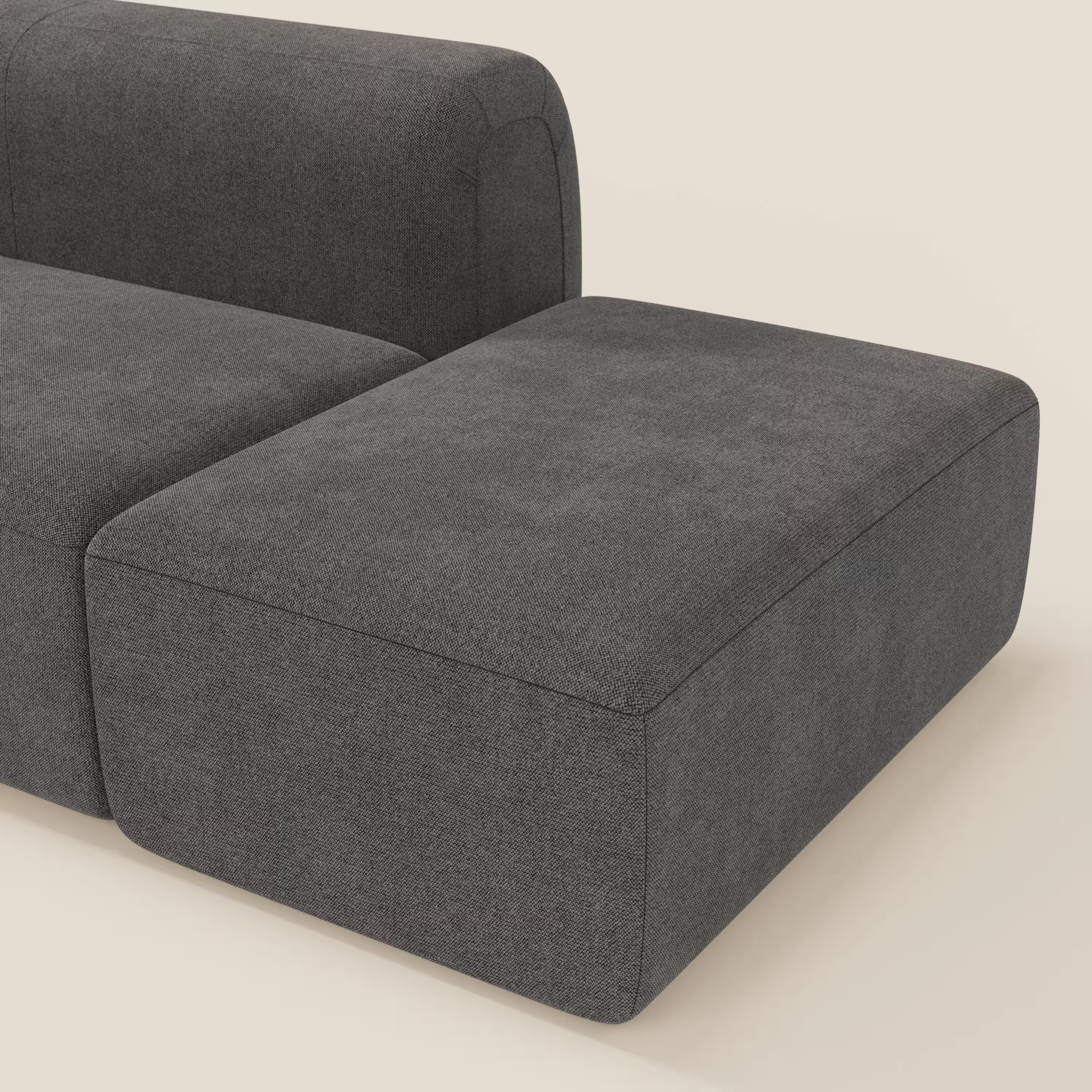 Primo piano di BonBon Divano componibile di Divani.Store in T23 antracite, che mostra l'elegante schienale, la seduta e l'ampio cuscino senza braccioli con pouf terminale su uno sfondo bianco sporco.