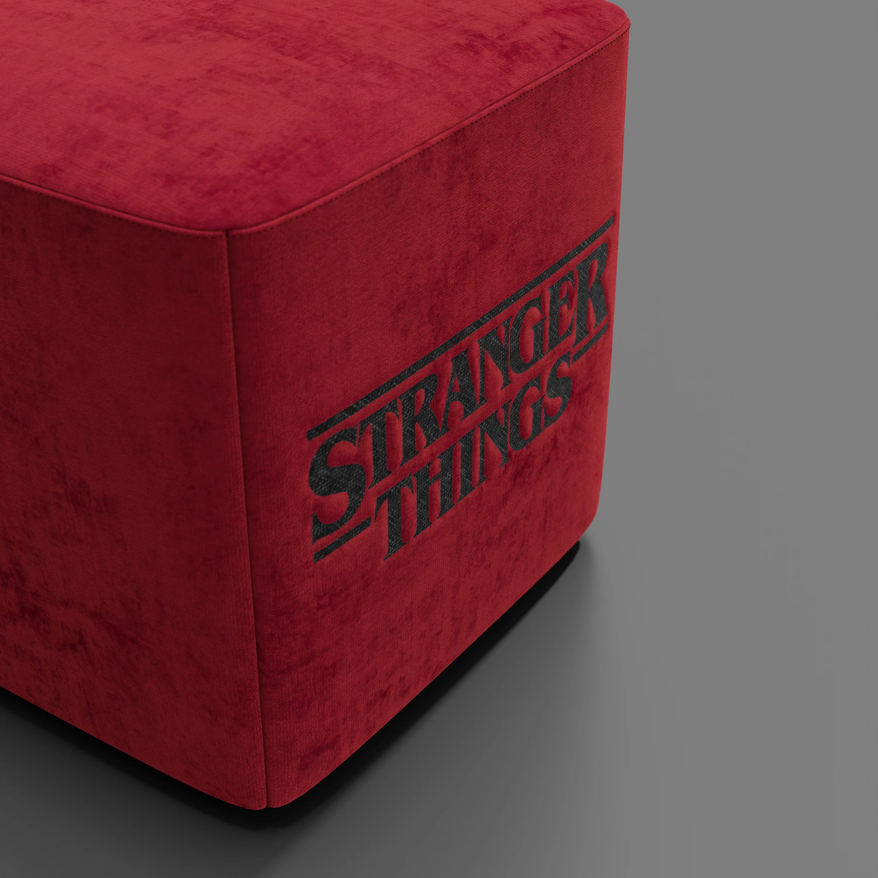 Il pouf Divani.Store Cube Stranger Things Edition, in tessuto rosso antimacchia con il logo nero della serie su un lato, è appoggiato su una superficie grigia liscia su uno sfondo grigio semplice.
