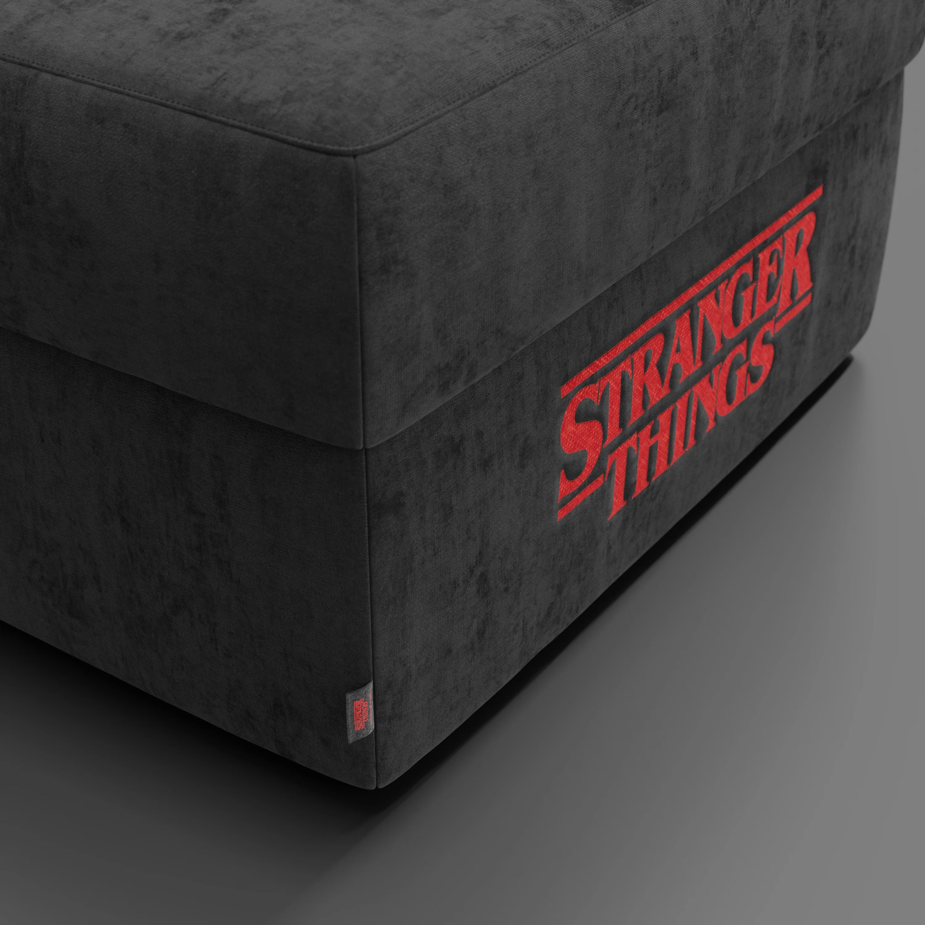 Un primo piano del pouf Divani.Store Sun Contenitore Stranger Things Edition - 60x80 cm in tessuto antracite lavabile - mostra il logo "Stranger Things" in rosso, la texture vellutata e una piccola etichetta vicino al bordo inferiore.