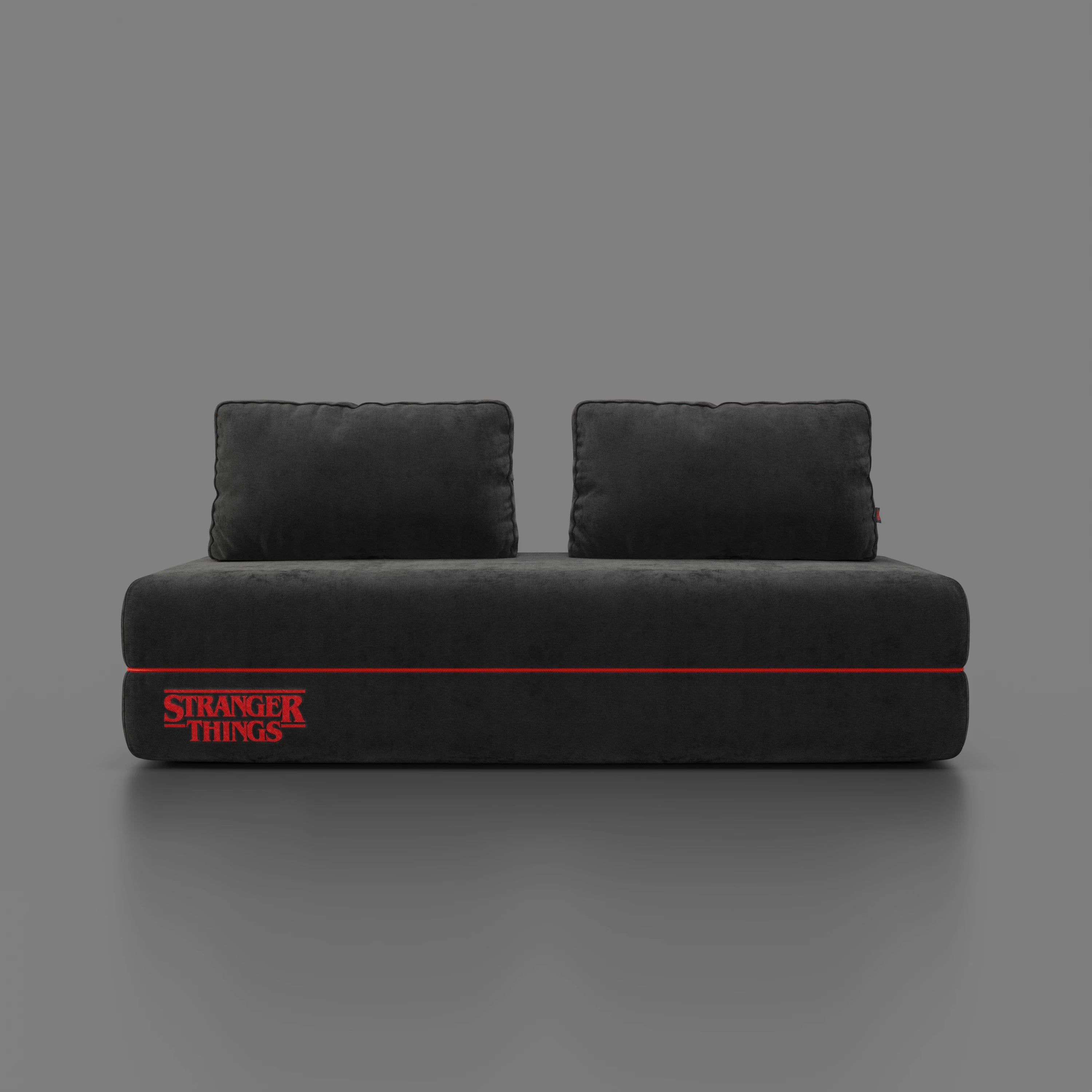 Un moderno divano letto matrimoniale Rift grigio scuro STRANGER THINGS Edition di Divani.Store con schienali mobili, striscia rossa e logo "Stranger Things" ricamato sul pannello frontale si staglia su uno sfondo grigio semplice.