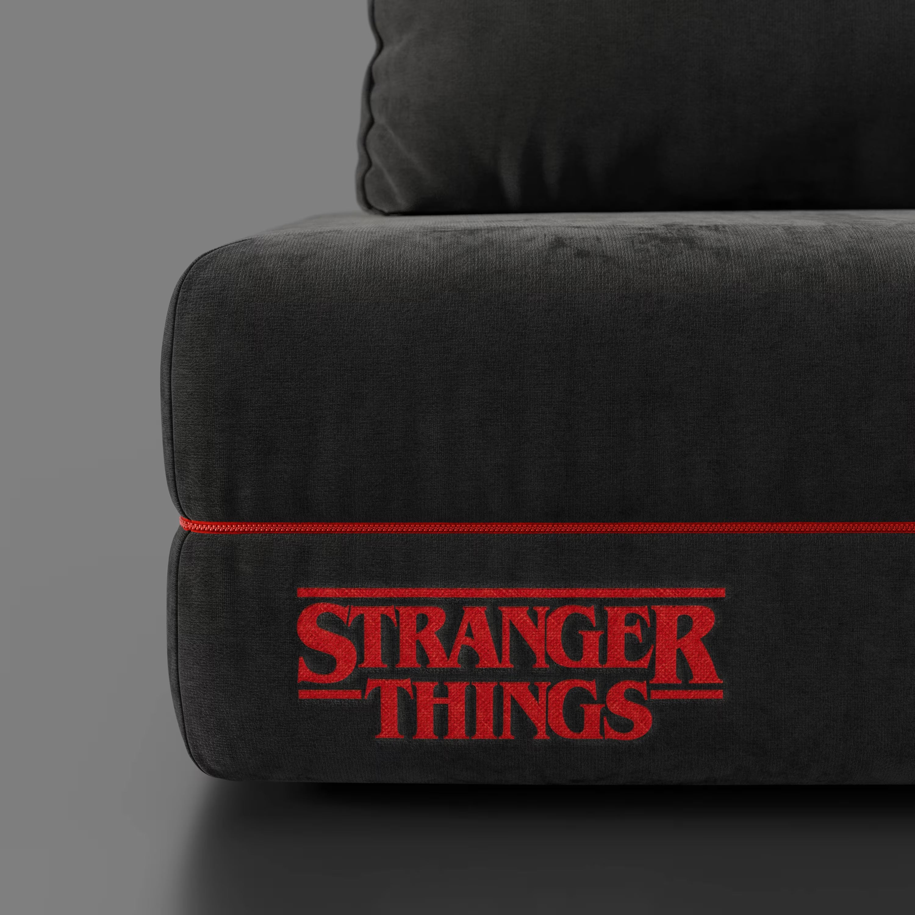 Primo piano del divano letto matrimoniale Rift STRANGER THINGS Edition di Divani.Store, caratterizzato da un tessuto antracite antimacchia, dal ricamo del logo "Stranger Things" in rosso e da una vistosa cucitura rossa su uno sfondo grigio minimal.