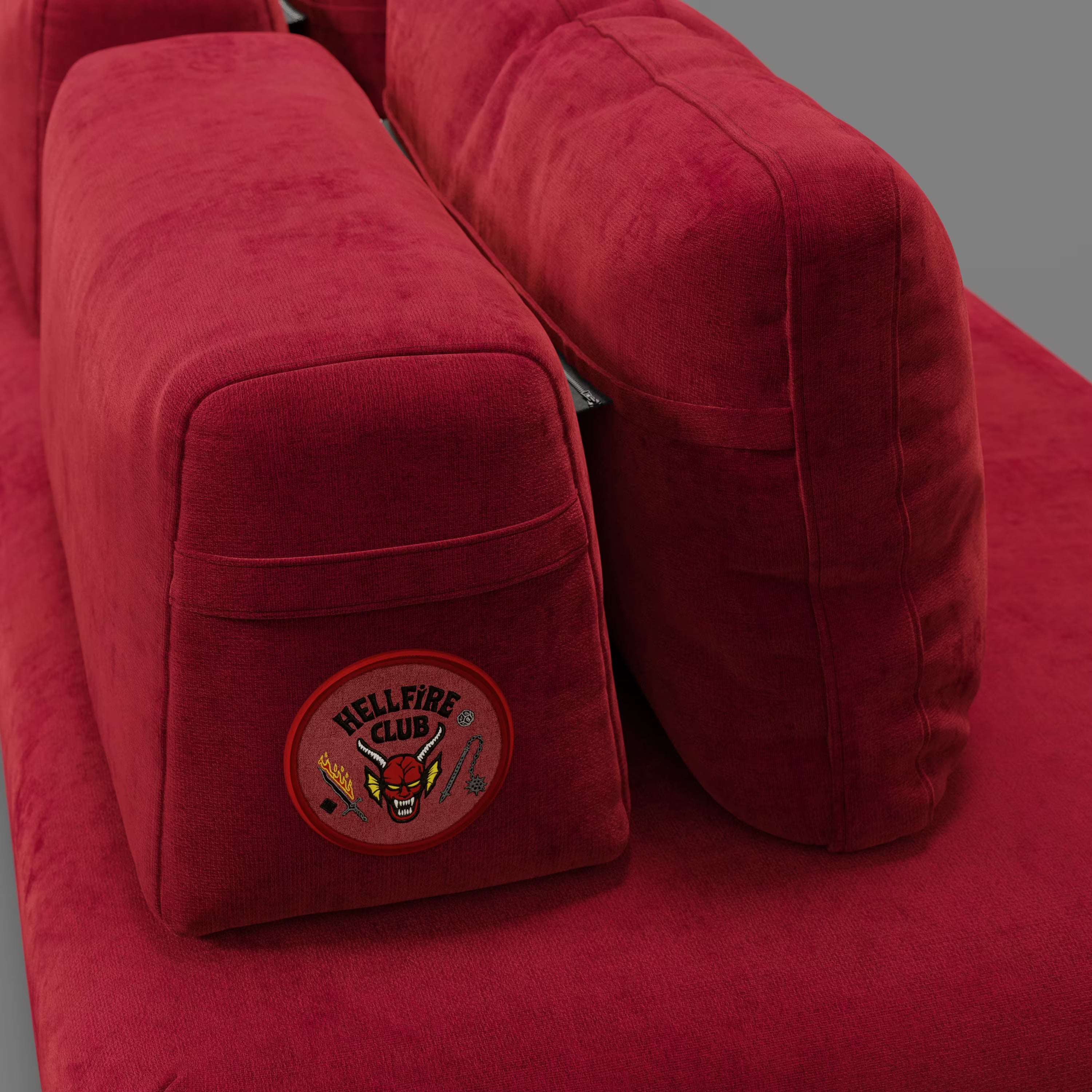 Primo piano del divano letto matrimoniale Rift STRANGER THINGS Edition di Divani.Store in tessuto rosso smacchiabile, caratterizzato da cuscini rettangolari, uno dei quali con patch "Hellfire Club" raffigurante un volto di diavolo e armi incrociate, su sfondo grigio.