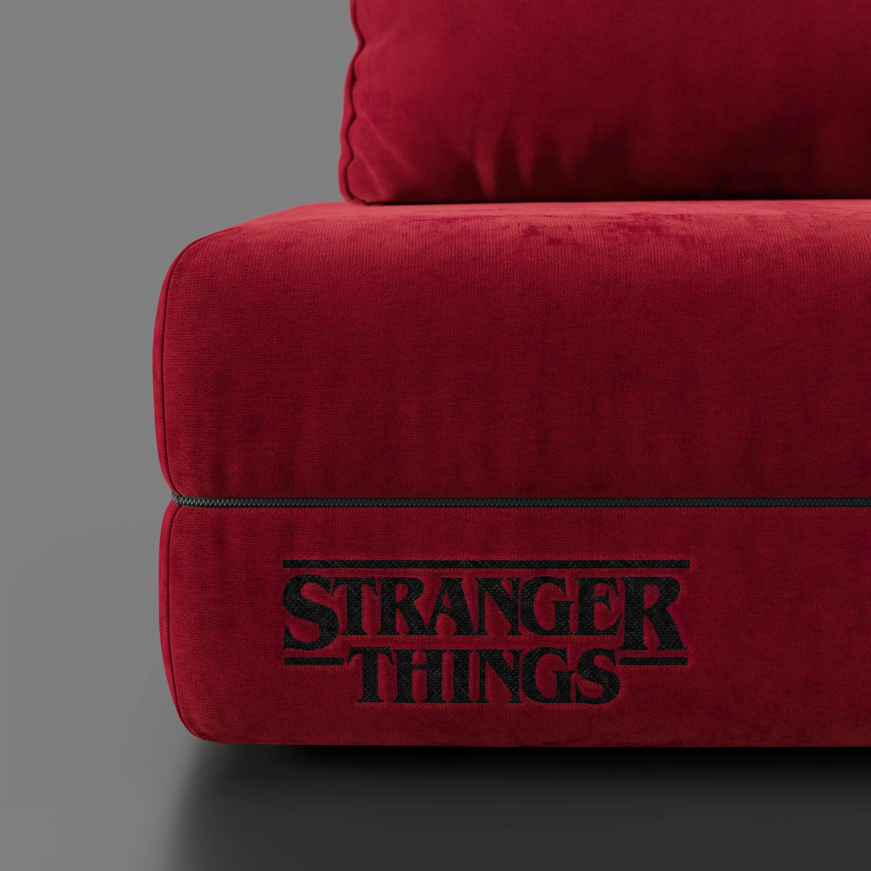 Un primo piano del divano letto matrimoniale Rift rosso STRANGER THINGS Edition di Divani.Store mostra un testo ricamato in nero su un tessuto morbido e antimacchia, ideale per gli appassionati. La seduta e gli schienali mobili spiccano su uno sfondo grigio.