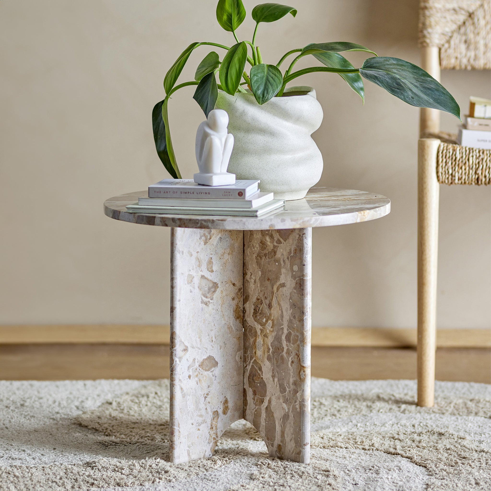 Un piccolo Jasmia Tavolino rotondo in marmo marrone di Divani.Store presenta una base beige testurizzata ed è sormontato da una pila di libri e una scultura astratta bianca. Un grande vaso in ceramica con piante verdi a foglia completa il design nordico, mentre è appoggiato su un leggero tappeto di peluche contro una parete beige.