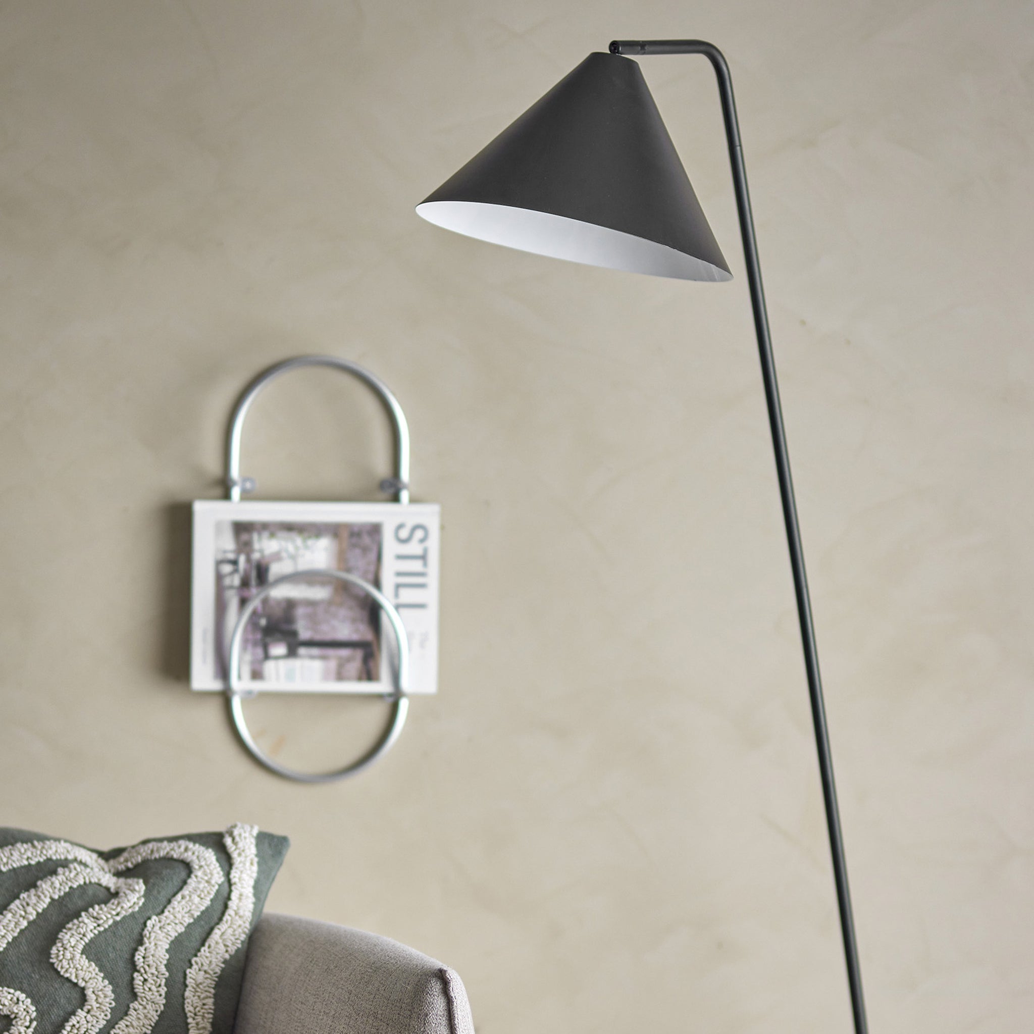 L'interno minimalista mette in mostra la lampada da terra dallo stile nordico Latisha in metallo nero di Divani.Store, caratterizzata da un elegante paralume conico. Un divano grigio è parzialmente visibile, decorato con un cuscino fantasia, mentre la parete espone un porta riviste montato che mostra "STILL", incarnando il moderno minimalismo nordico.