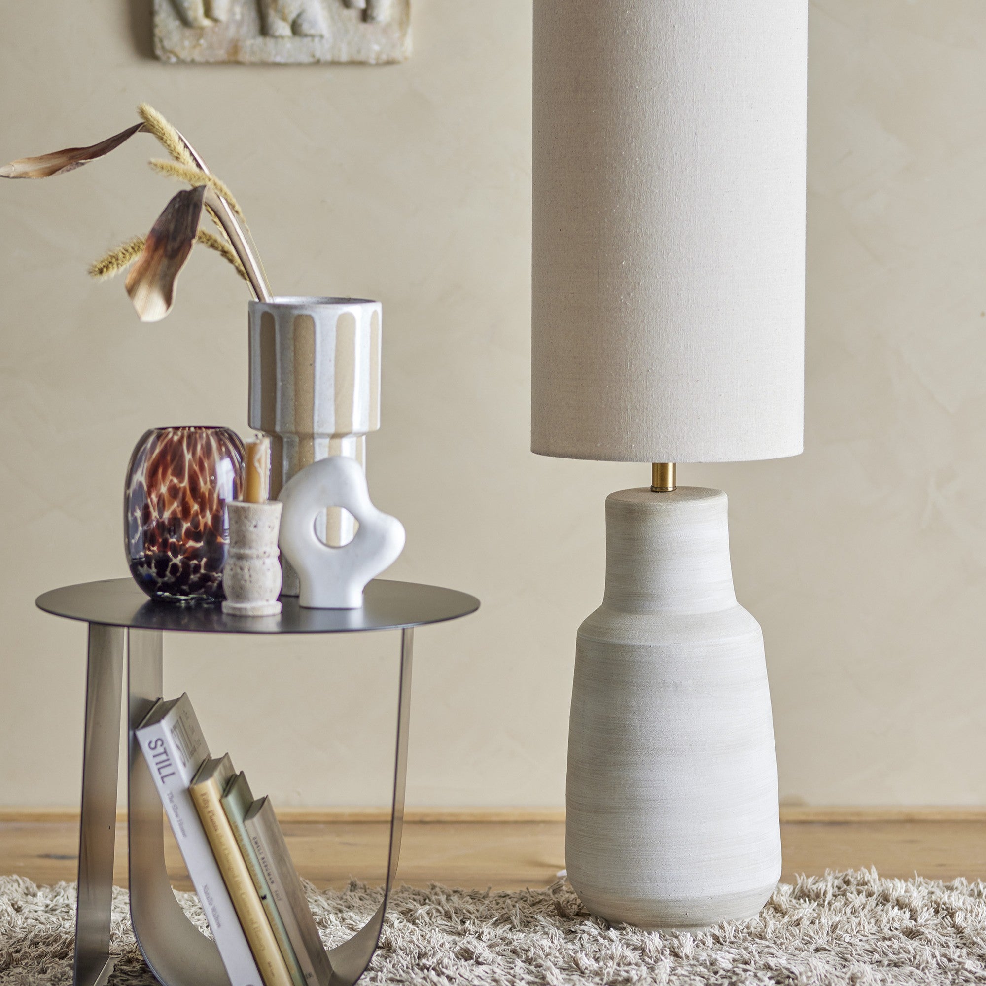 Un tavolino rotondo con base in metallo sorregge vasi decorativi e fiori secchi. Nelle vicinanze, la lampada da terra Linetta con base in gres stoneware bianco di Divani.Store, con paralume beige, è appoggiata su un tappeto texturizzato. L'arredamento sofisticato e minimalista con toni neutri crea un'atmosfera accogliente e moderna. I libri sono infilati nella curvatura della base del tavolo.