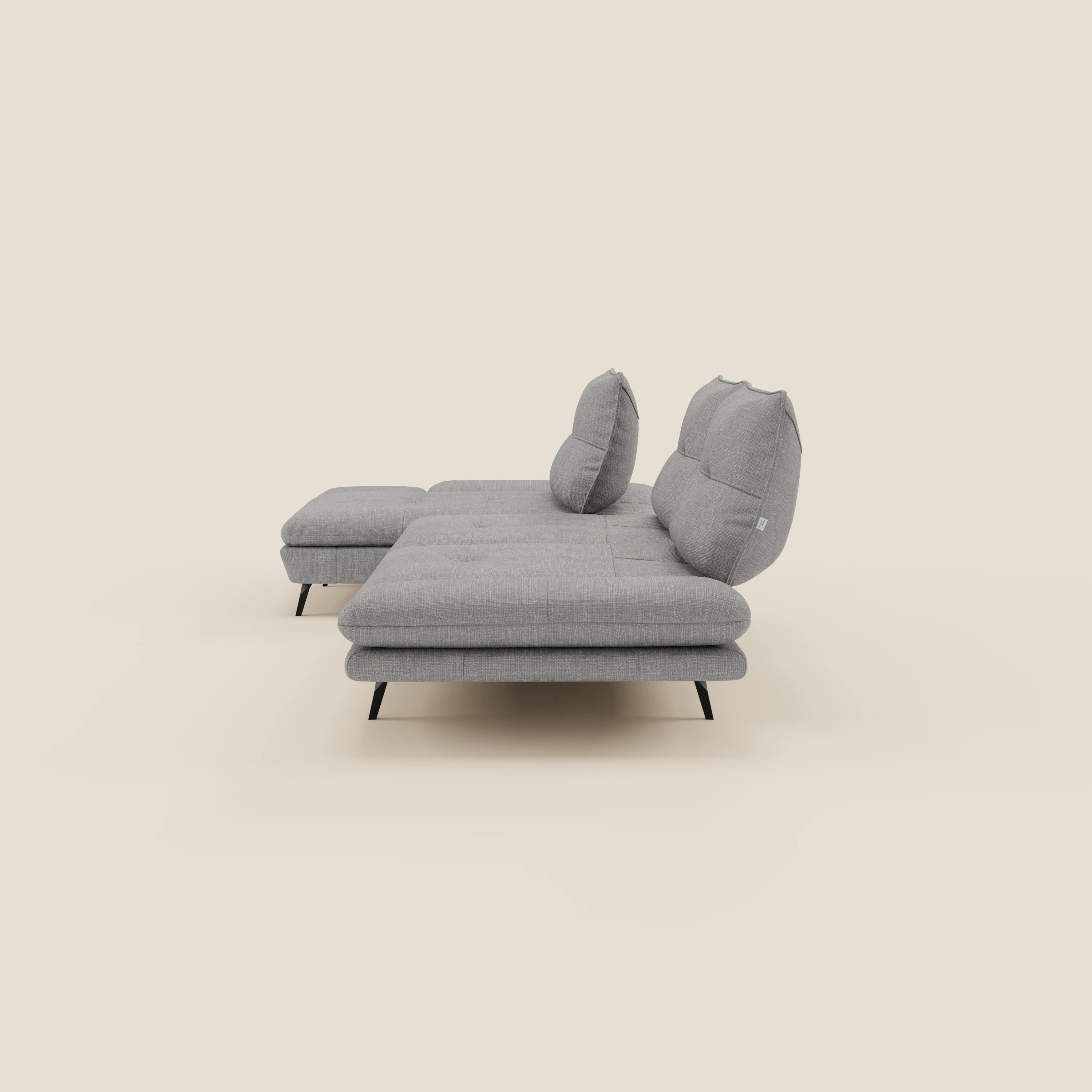 Il Divano con penisola Ardex di Divani.Store, rivestito in elegante tessuto impermeabile grigio chiaro T06 con schienali traslanti e braccioli regolabili, è mostrato di profilo con il pouf abbinato a sinistra su uno sfondo crema.