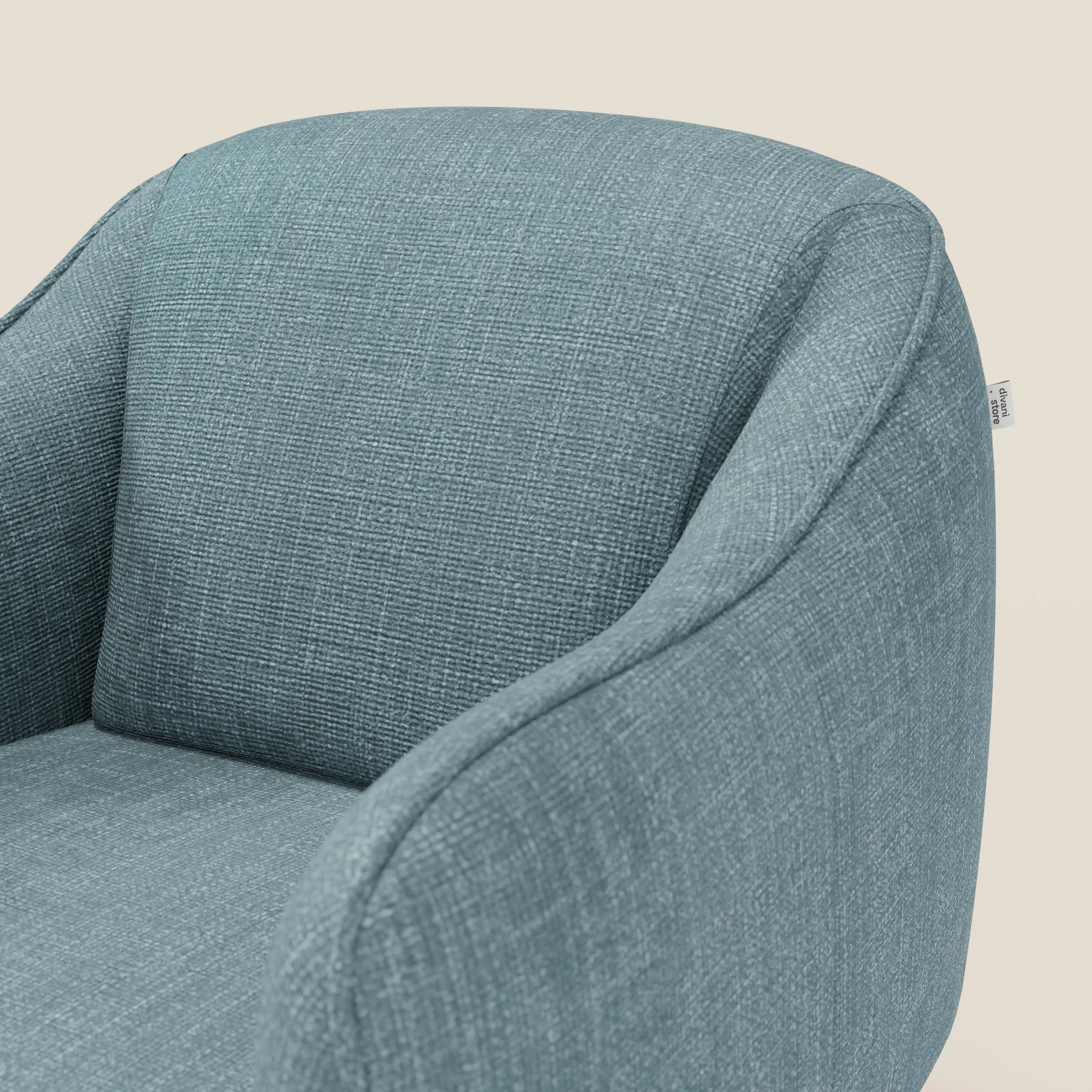 Primo piano di Avela Poltroncina di Divani.Store, rivestita in tessuto impermeabile T06 azzurro, con schienale curvo e braccioli arrotondati. Una piccola etichetta laterale in tessuto spicca sullo sfondo chiaro.