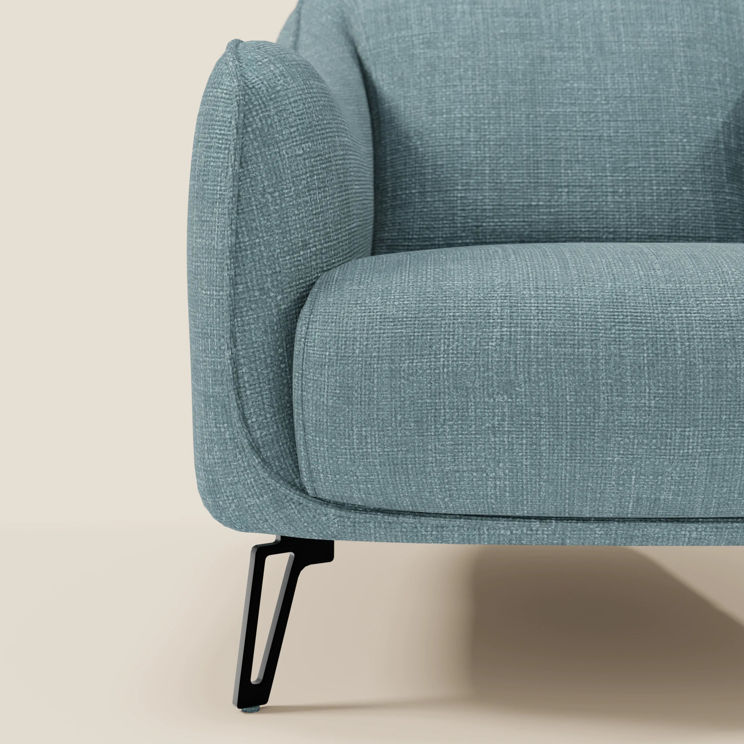 Primo piano di Avela Poltroncina di Divani.Store in morbido tessuto azzurro T06 impermeabile. L'immagine mostra il bracciolo curvo, la seduta imbottita e la gamba angolata in metallo nero su uno sfondo beige semplice per un look elegante.