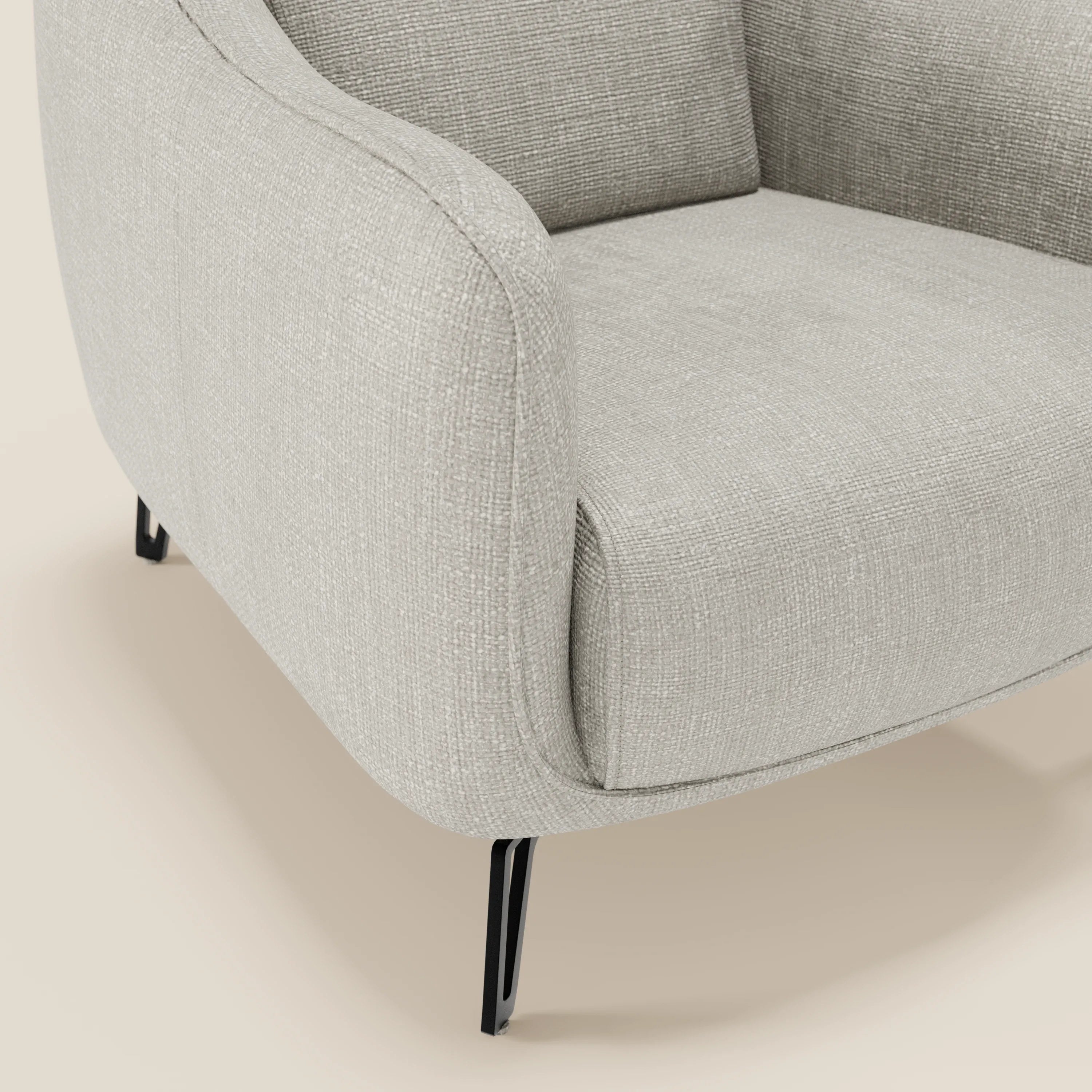 Primo piano di Avela Poltroncina di Divani.Store, caratterizzata da un comfort senza tempo in morbido tessuto grigio chiaro T06 resistente all'acqua, braccioli delicatamente curvati e sottili gambe in metallo nero su uno sfondo beige chiaro.