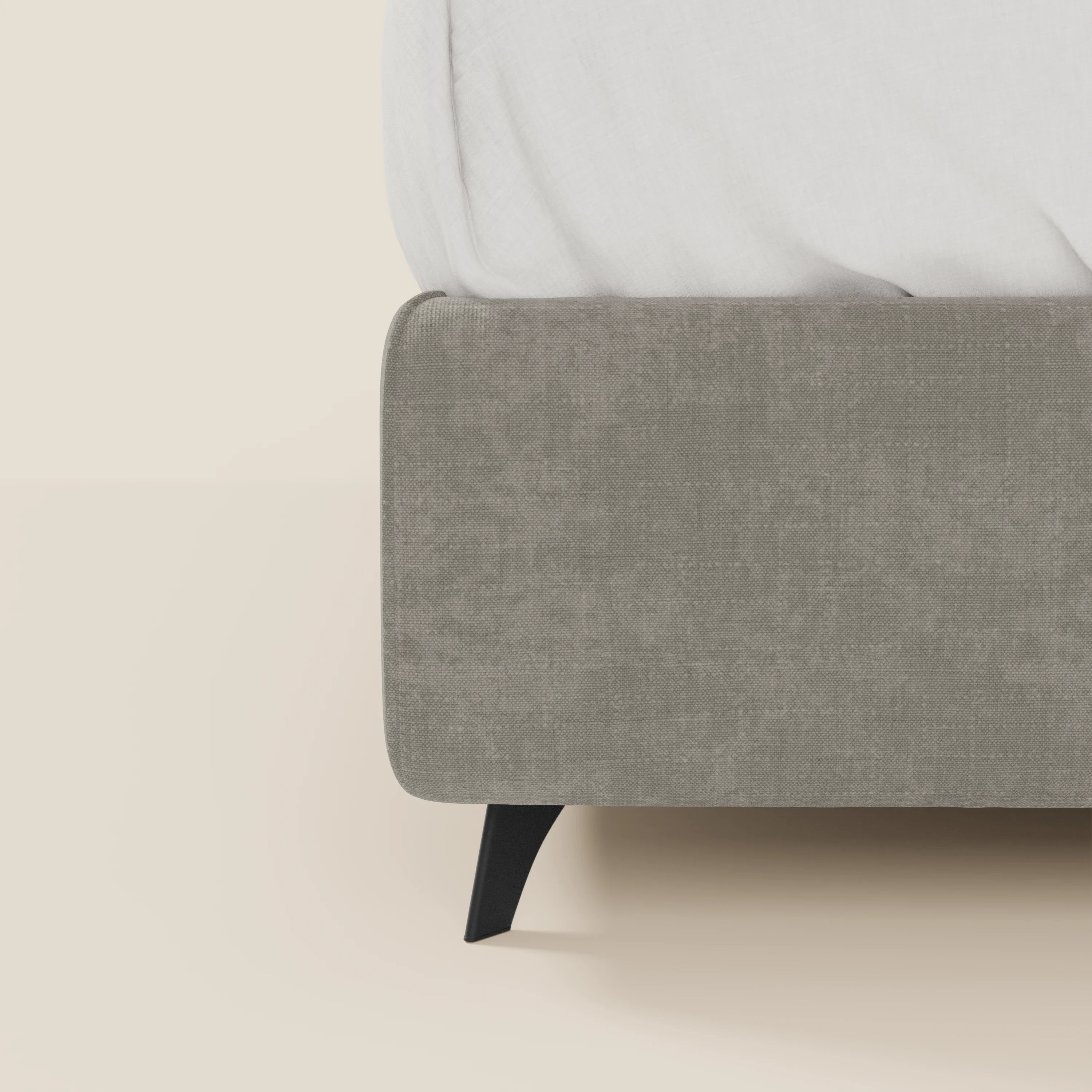 Primo piano del letto contenitore Bogart Edizione Grande Fratello di Letti.store, in tessuto T22 antimacchia grigio chiaro, sfoderabile e con gamba angolare nera. Il letto e il materasso sono visibili solo in parte, la biancheria è bianca su sfondo beige.