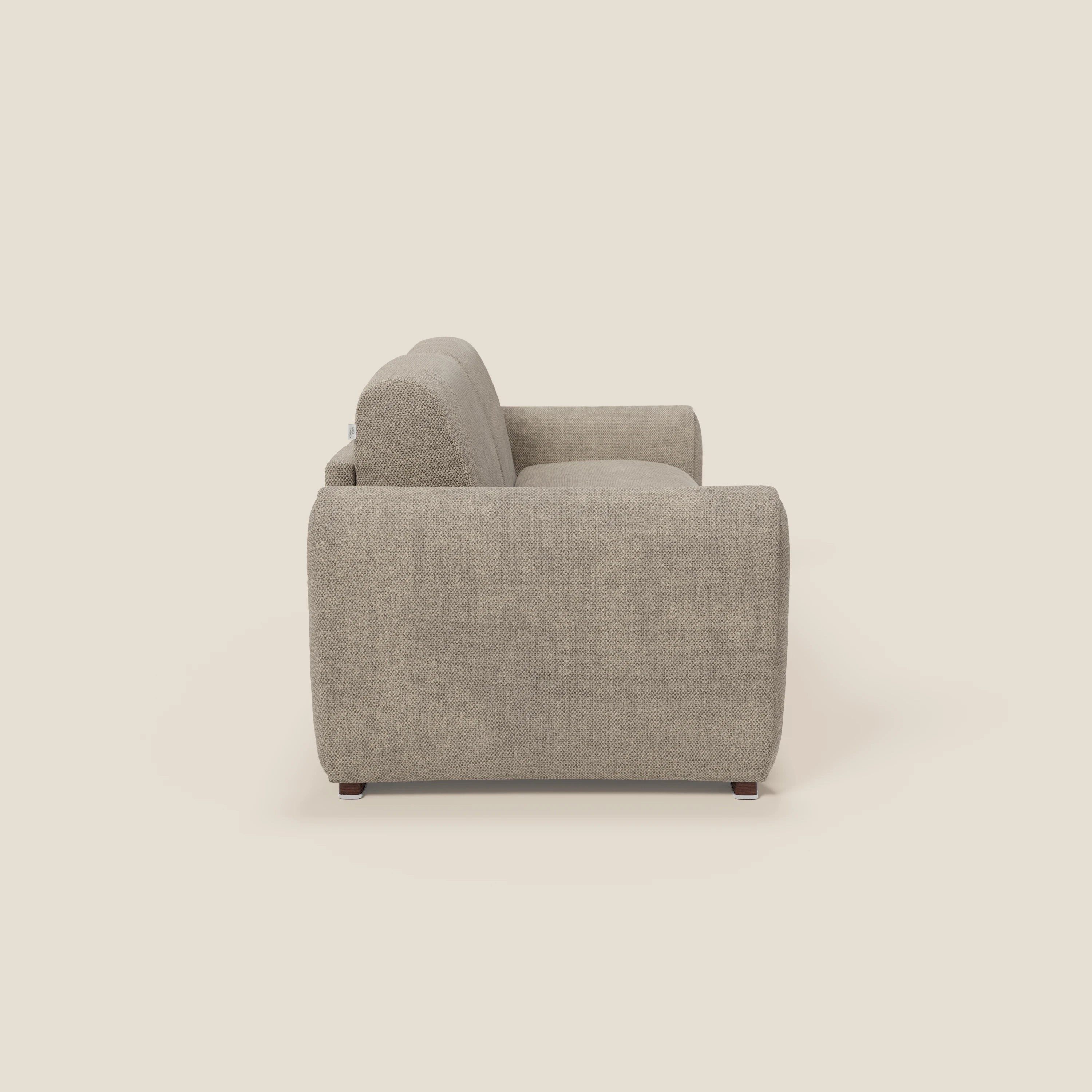 Una vista laterale del divano letto moderno Kaleo di Divani.Store in elegante tessuto impermeabile T26 tortora, caratterizzato da bordi arrotondati, ampi braccioli, seduta e schienale morbidi, si staglia su uno sfondo bianco sporco.