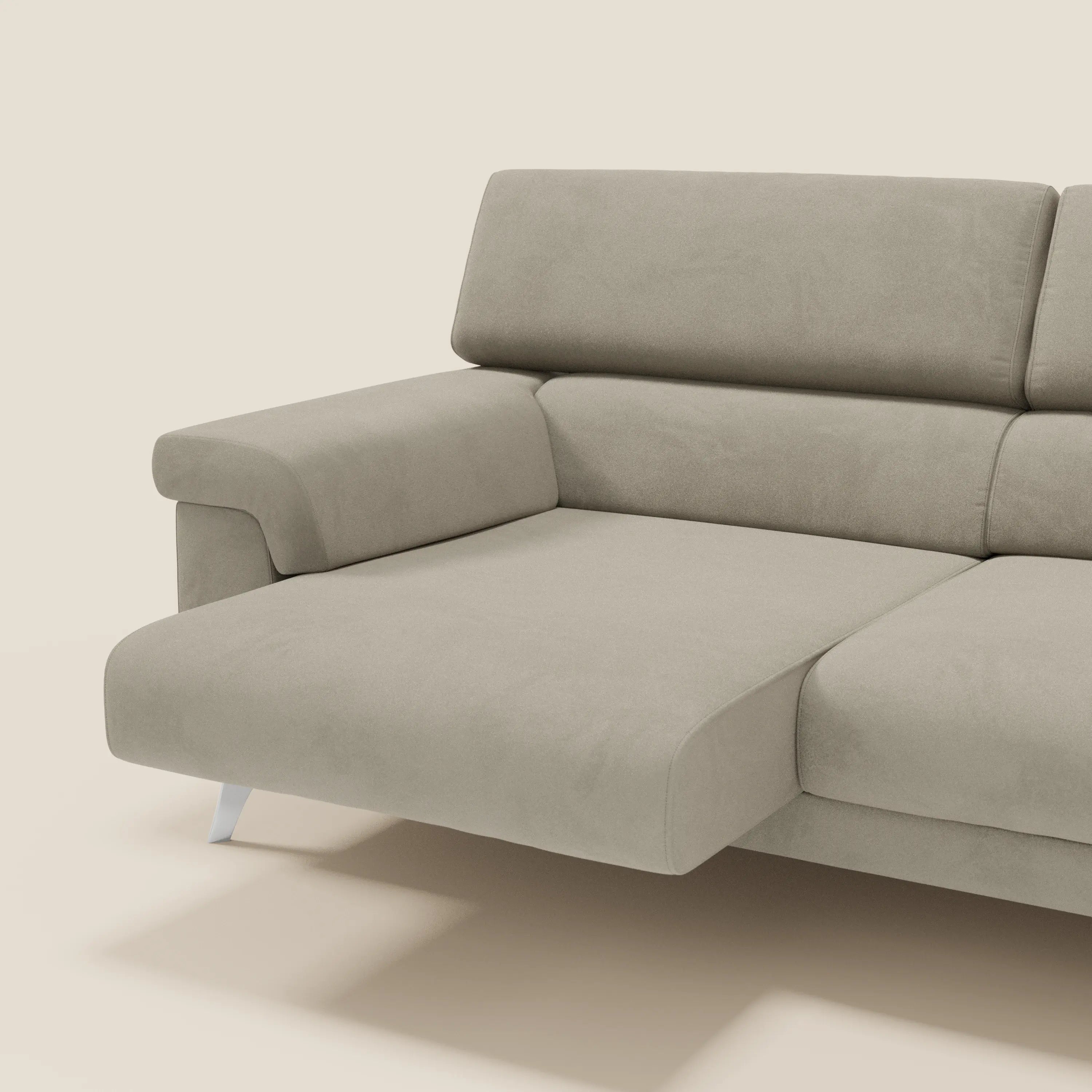 Un primo piano del divano Raffaello di Divani.Store, in smacchiabile microfibra tortora T11, mostra le sedute allungabili e i poggiatesta reclinabili. La seduta di sinistra è estesa come una chaise longue, con ampi braccioli, cuscini in feltro e corte gambe metalliche su uno sfondo beige.